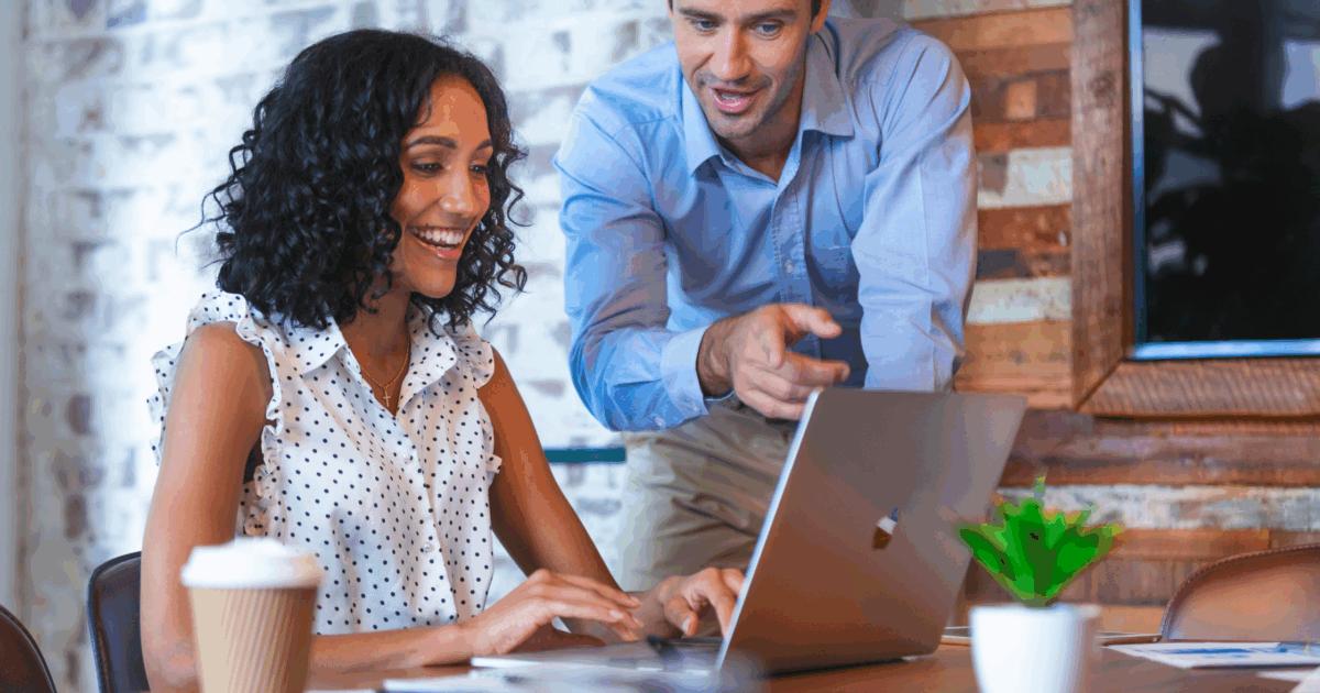 Female sitting at table, smiling, using laptop while male stands next to her pointing out something on her screen