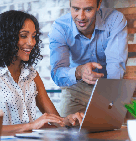 Female sitting at table, smiling, using laptop while male stands next to her pointing out something on her screen