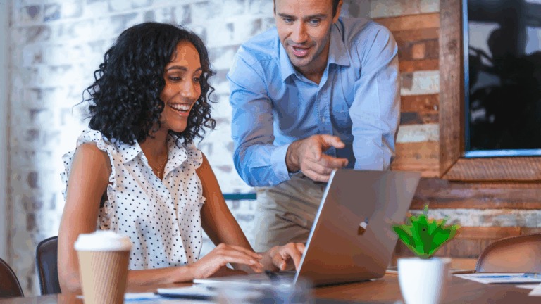 Female sitting at table, smiling, using laptop while male stands next to her pointing out something on her screen