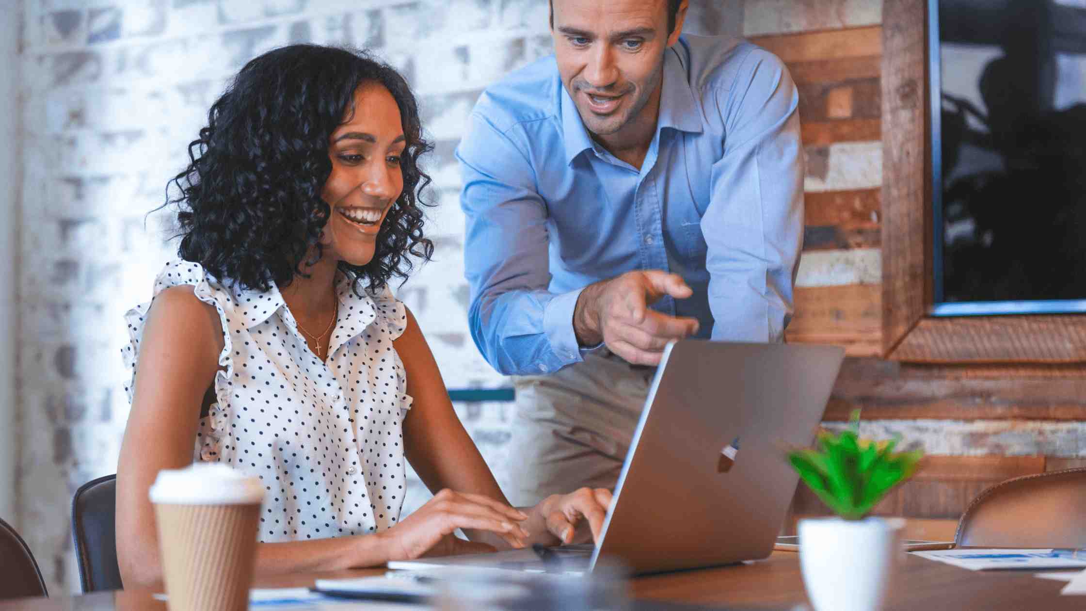Female sitting at table, smiling, using laptop while male stands next to her pointing out something on her screen