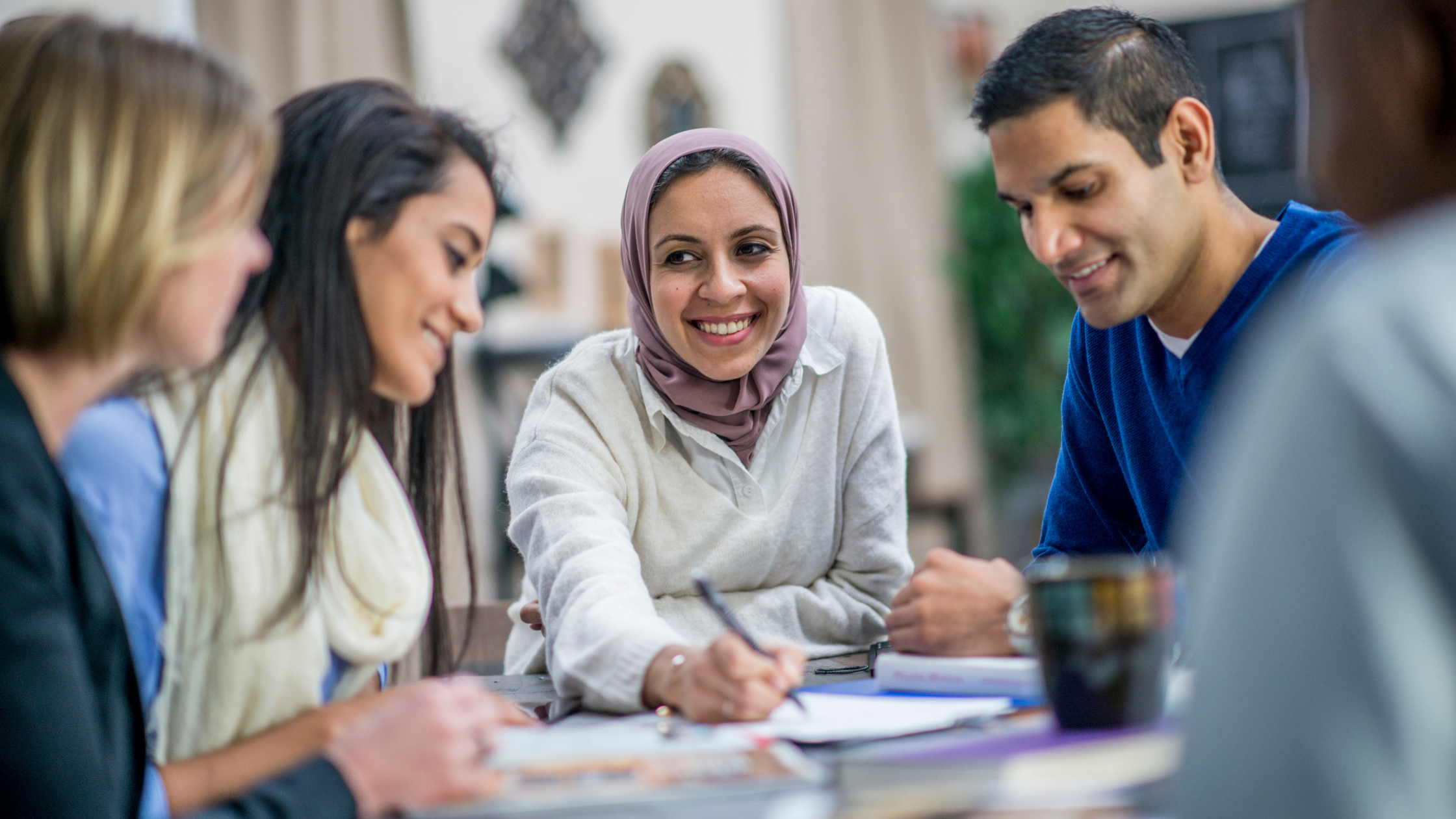 Photo of 5 people gathered around a table collaborating on a project while looking at paper on the table