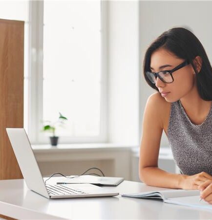 Female looking at laptop screen, taking notes in a notebook