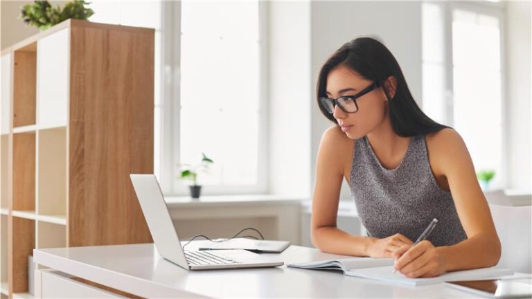 Female looking at laptop screen, taking notes in a notebook