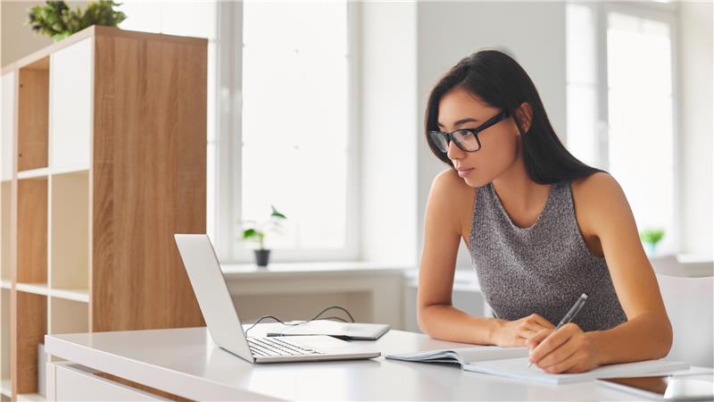 Female looking at laptop screen, taking notes in a notebook
