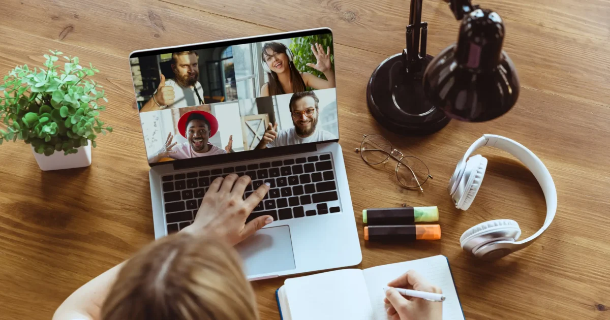 female taking notes on a team virtual meeting, members displayed on laptop screen
