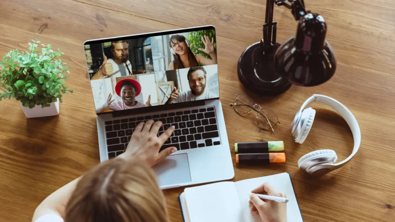 female taking notes on a team virtual meeting, members displayed on laptop screen