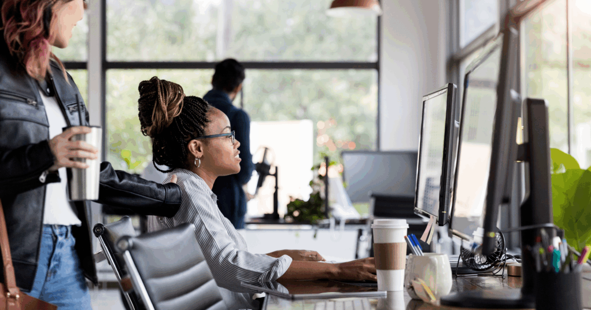 Female working on computer at her desk in offer. Coworker is seen engaging with her.