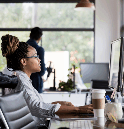 Female working on computer at her desk in offer. Coworker is seen engaging with her.