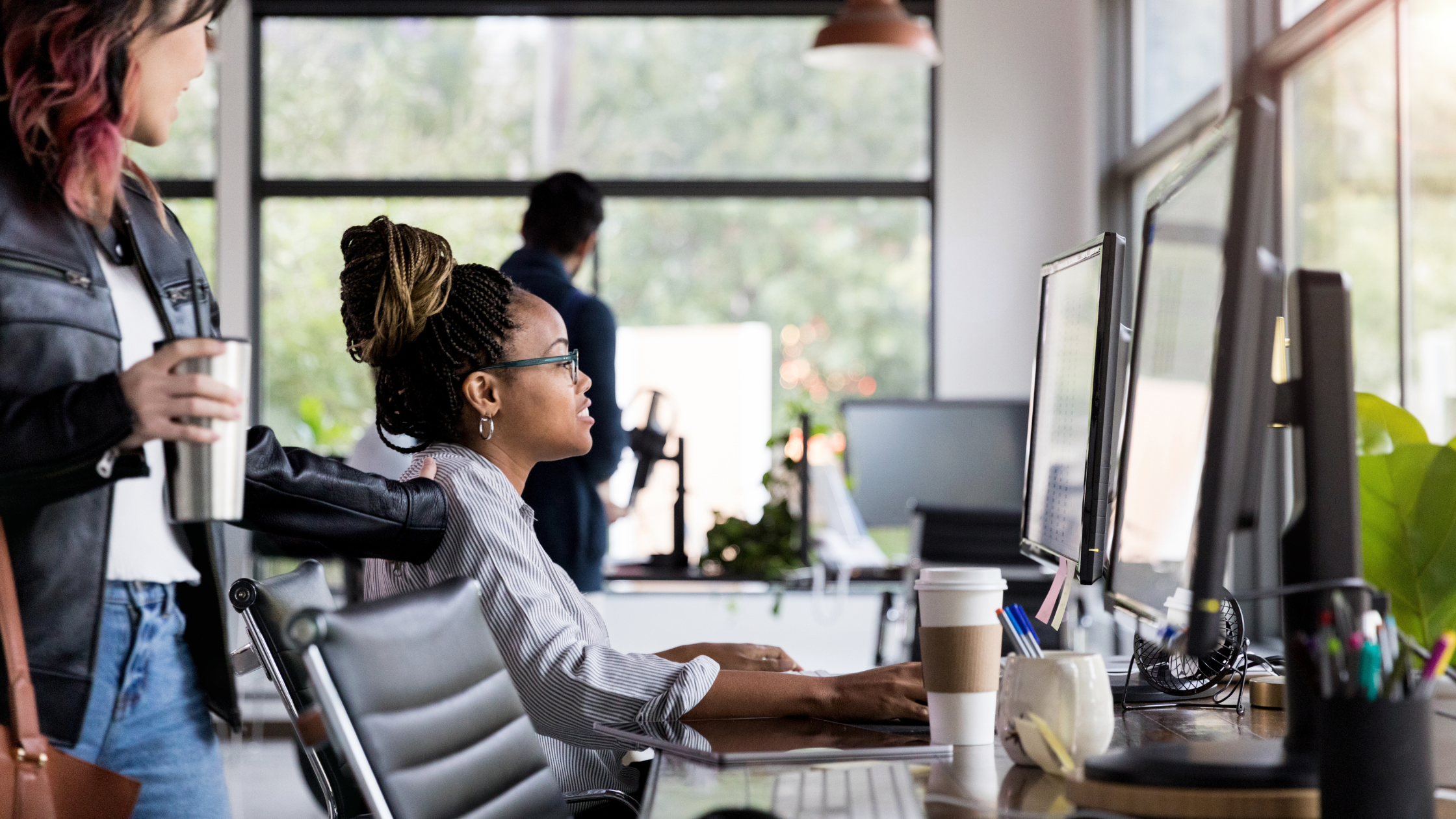 Female working on computer at her desk in offer. Coworker is seen engaging with her.
