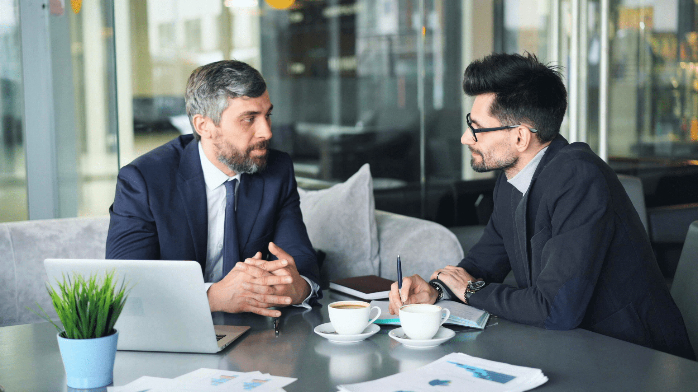 Two male coworkers talking at a table equipped with a laptop and coffees