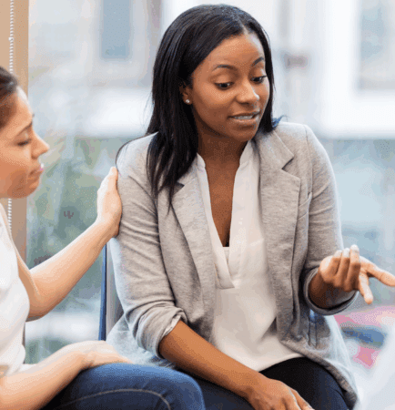 Female seated, talking to group of people. Another female is showing connection on her face and embracing her arm.